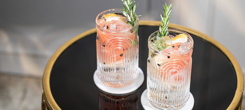 Two ribbed crystal tumblers of a pale pink drink over ice, garnished with rosemary and a wheel of citrus, on a dark mirrored tray