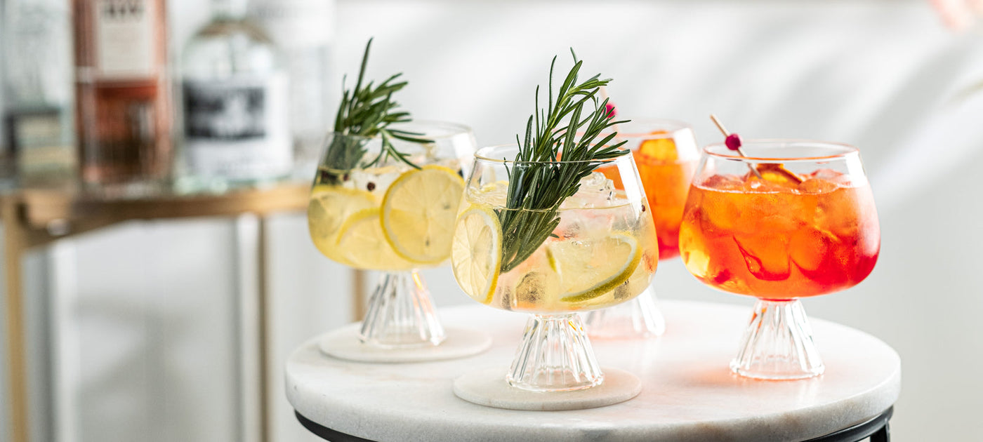 Tre calici in cristallo con aperitivi analcolici su un piano di marmo, guarniti con rosmarino, una fetta di limone e una di arancia