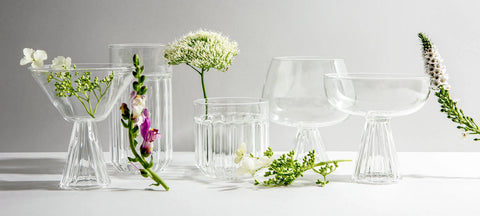 A row of clear crystal glasses, a martini glass, a highball, a lowball, a Nick and Nora and a coupe, with fresh flowers