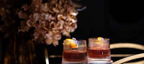 Two ribbed lowball glasses with an amber cocktail on a gold bar cart beside dried hydrangeas