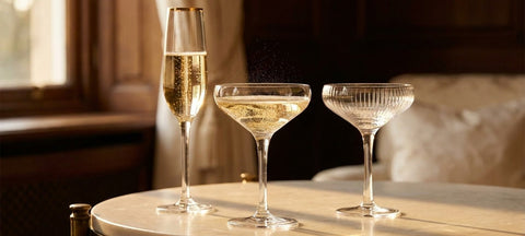 Coppa da champagne in cristallo con Champagne dorato accanto a una coppa Art Deco e a un flute su marmo chiaro