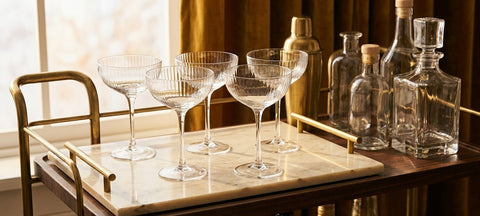 Four empty ribbed champagne coupes on a marble tray of a brass bar cart beside a shaker and unlabeled bottles in winter evening light