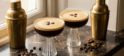 Two espresso martinis with thick crema and coffee beans in crystal coupe glasses between brass cocktail shakers on dark marble