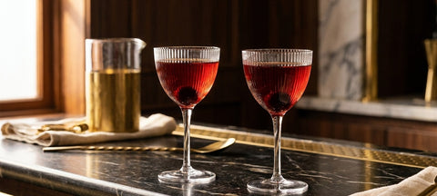 Two Manhattan cocktails with a cherry in ribbed Nick and Nora glasses on a dark marble bar beside a brass mixing glass