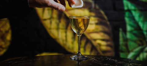A hand garnishing a stirred cocktail in a Nick and Nora glass against a painted tropical wall