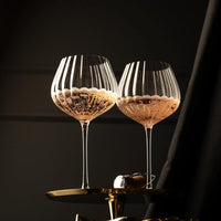 Two elegant clear wine glasses with vertical fluting, partially filled with bubbly pink champagne, set on a gold-toned stand against a dark draped background.