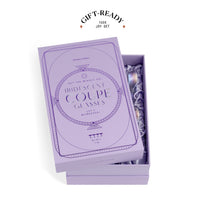 A lavender gift box set for George Clooney Iridescent Coupe Glasses, shown open with the box lid leaning on top and revealing packaged iridescent glasses inside. A 'GIFT-READY 100% JOY SET' badge is printed above the box on a clean white background.