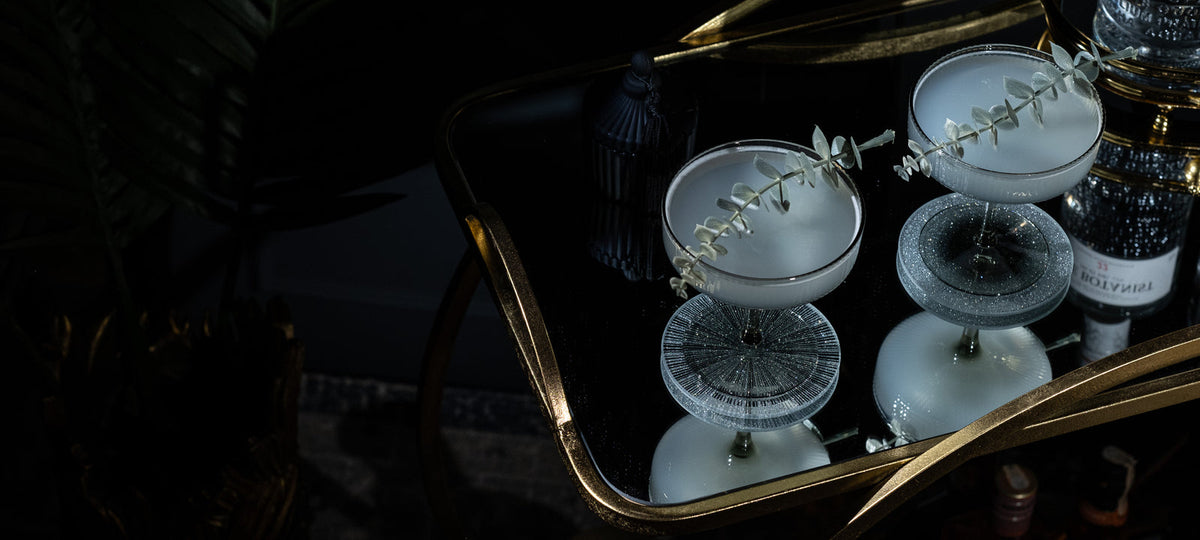 Smoke-black Art Deco coupes with eucalyptus on a dark gold bar cart in low speakeasy light