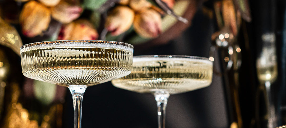 Crystal coupes with gold rims lined up on a brass bar cart in candlelight