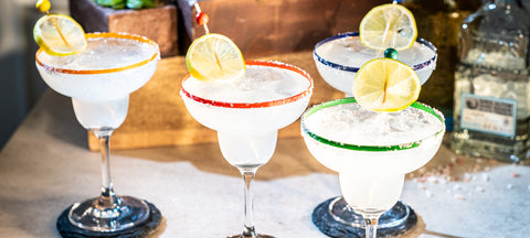Three margaritas with coloured salt rims and lime wheels in stemmed margarita glasses on a wooden crate