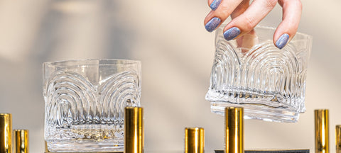 Two hand-blown crystal Art Deco lowball glasses, one lifted by hand, on a gold bar cart