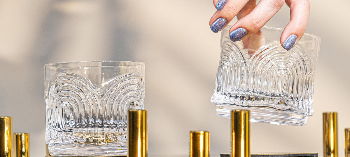 Two hand-blown crystal Art Deco lowball glasses, one lifted by hand, on a gold bar cart