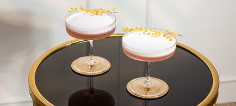 Two pink foam cocktails with yellow blossoms in ribbed coupes on a black and gold side table