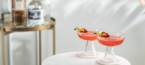 Two coupe cocktails with edible flowers on a marble side table beside a gold bar cart