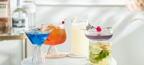 Four cocktail glass shapes side by side, a martini glass, a highball, a coupe and a lowball, each filled with a different drink