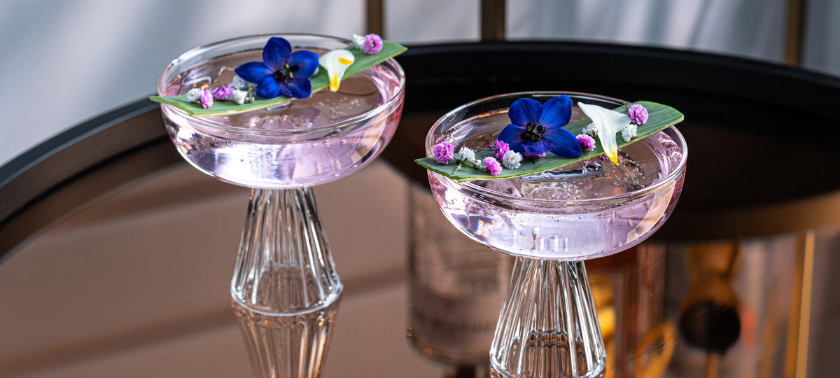 Two crystal coupes of a pale pink shaken mocktail with edible flowers on a mirrored bar cart