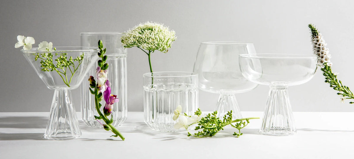 A row of clear crystal glasses, a martini glass, a highball, a lowball, a Nick and Nora and a coupe, with fresh flowers