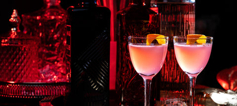 Two pink Cosmopolitans with an orange twist in stemmed Nick and Nora glasses under red bar light
