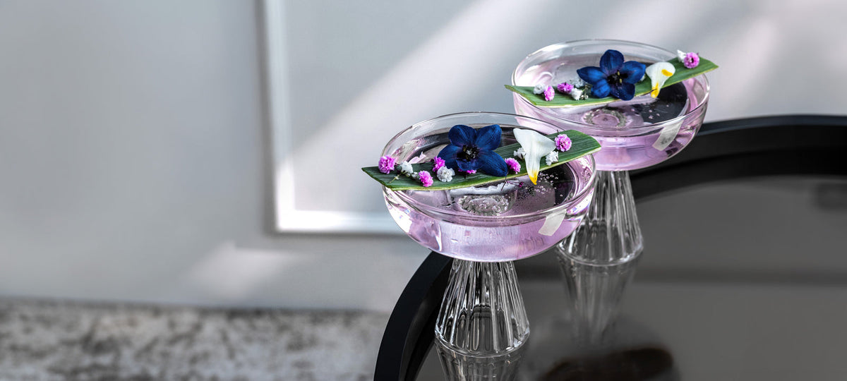 Two lilac cocktails with edible flowers in ribbed coupes on a black table