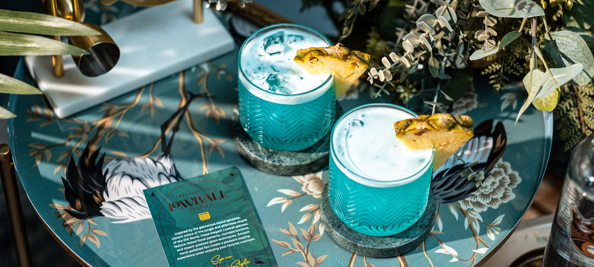Due tumbler bassi in cristallo rigato con drink analcolici azzurri su ghiaccio, guarniti con spicchi di ananas, su un vassoio verde acqua