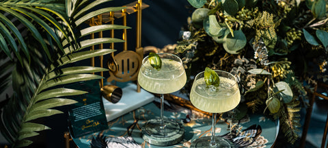 Two pale green gimlets with a basil leaf in ribbed crystal coupe glasses among palm leaves