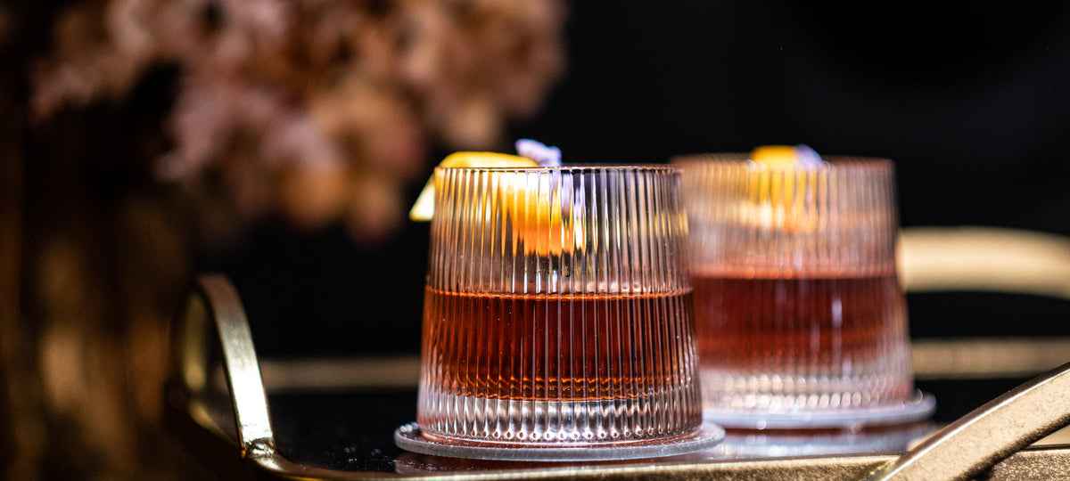 Two ribbed Art Deco lowball glasses with an amber cocktail and orange peel on a gold bar cart in low light