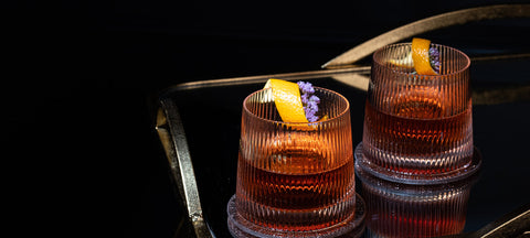 Zwei geriffelte Tumbler mit einem bernsteinfarbenen alkoholfreien Drink auf Eis, garniert mit Orangenzeste und Lavendel, auf einem dunklen Spiegeltablett