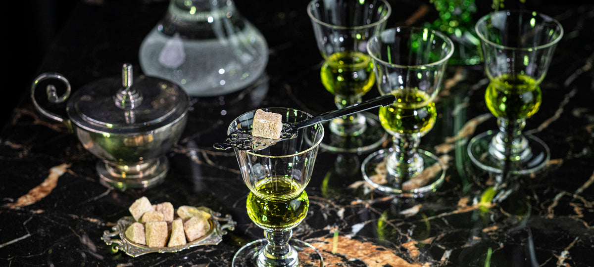 Un verre à absinthe Pontarlier avec une cuillère ajourée et un sucre, fontaine en arrière-plan sur marbre noir