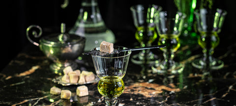 A Pontarlier absinthe glass with a slotted spoon and sugar cube beside a fountain and sugar cubes on black marble