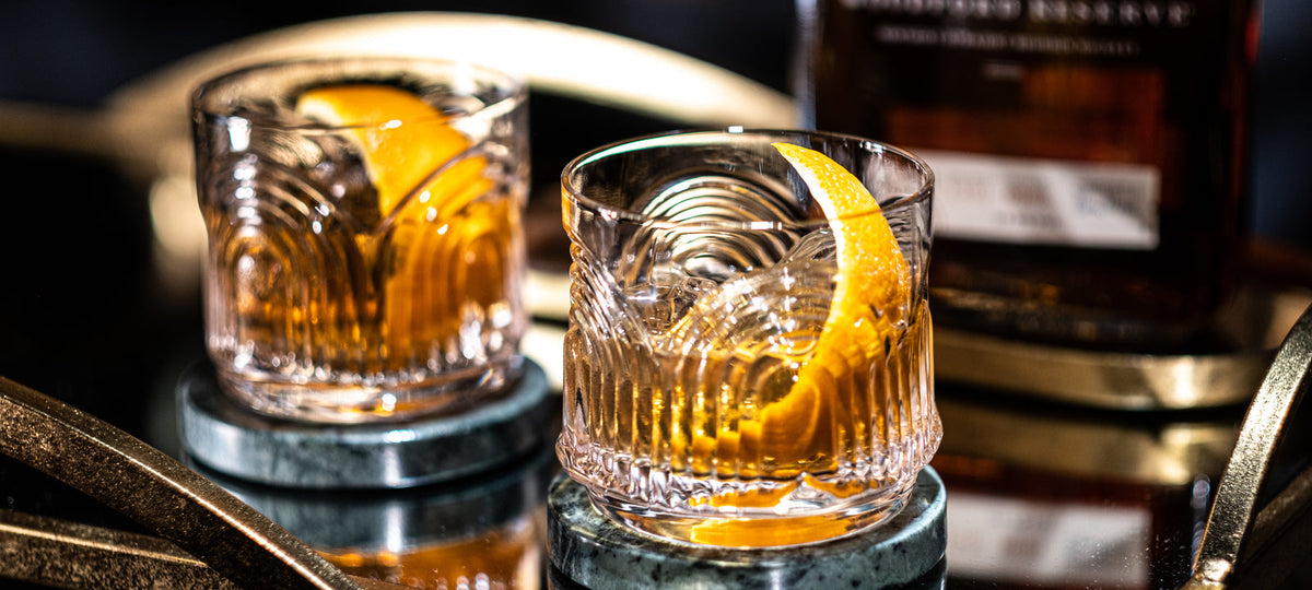 Two Art Deco rocks glasses with an Old Fashioned and a wide orange peel over ice on a gold bar cart