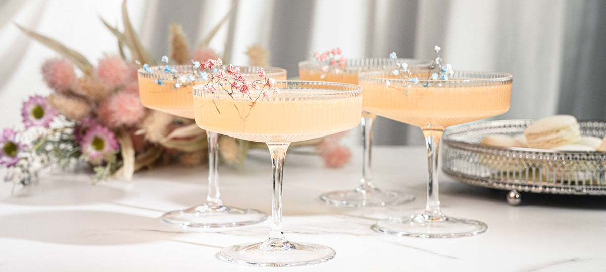 Three ribbed crystal coupes with a peach cocktail on a white table with dried flowers and macarons