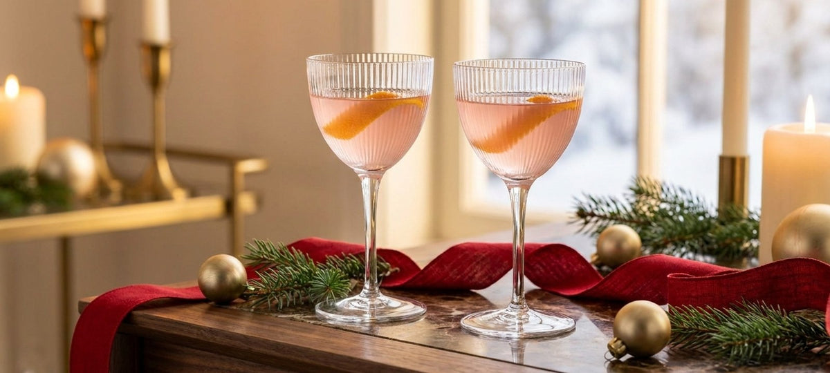 Dos copas Nick and Nora con un cóctel rosa y piel de naranja sobre una mesa de madera con lazo rojo, ramas de abeto y bolas doradas