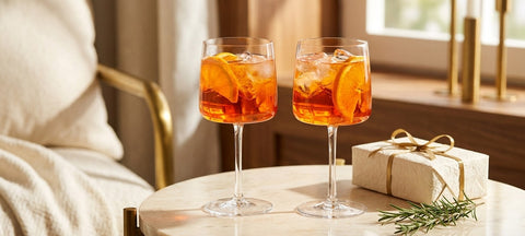 Two Aperol spritz cocktails with orange slices in stemmed crystal glasses on a marble side table beside a small wrapped gift and rosemary
