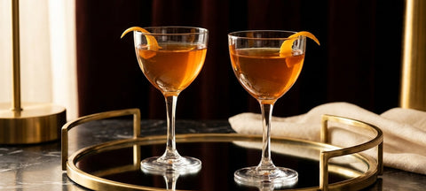 Two Nick and Nora glasses with amber stirred cocktails and an orange twist on a mirrored brass tray
