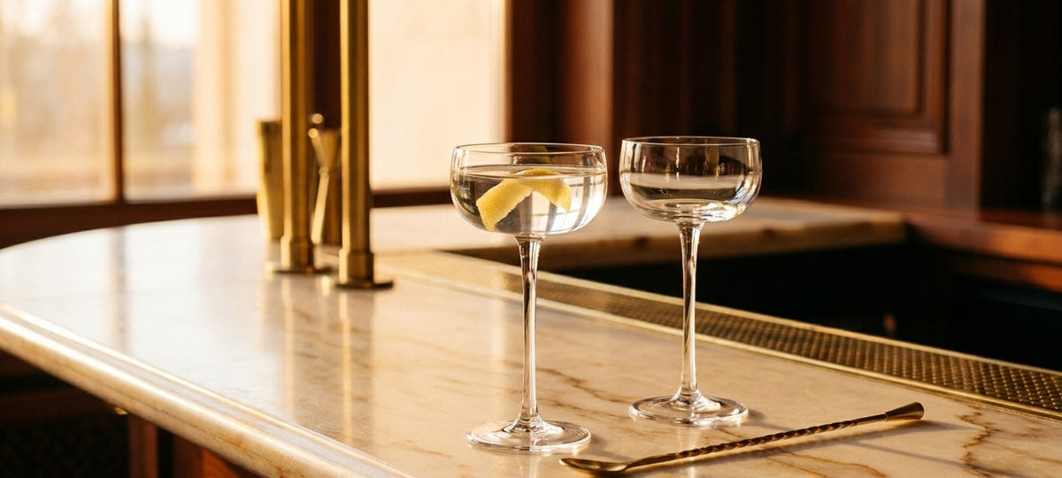 Bicchiere Nick & Nora in cristallo con un Dry Martini e scorza di limone accanto a una coppa Art Déco su un bancone di marmo chiaro