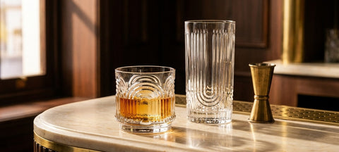 Bicchiere tumbler basso in cristallo a coste con un cubetto di ghiaccio accanto a un highball su un bancone di marmo chiaro