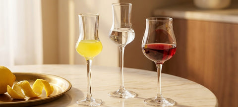 Bicchiere da liquore sfaccettato con limoncello accanto a un bicchiere da grappa a stelo lungo e a una copita da porto su marmo chiaro