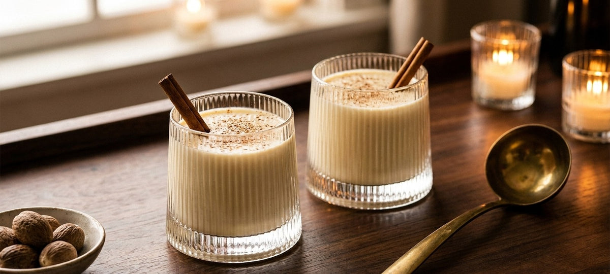 Two ribbed lowball glasses of eggnog dusted with nutmeg with cinnamon sticks on a dark wood table beside a brass ladle and candles