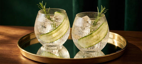 Zwei gerillte Art-Deco-Gin-Ballonglaeser mit Gin Tonic, Gurkenscheibe und Rosmarin auf einem Messingtablett