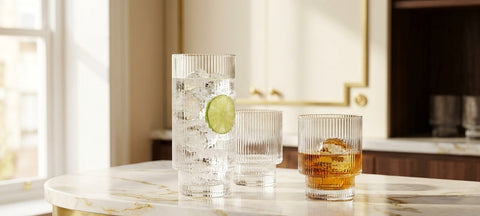 A tall ribbed highball glass with gin and tonic beside a ribbed Art Deco lowball glass with whiskey on a marble counter