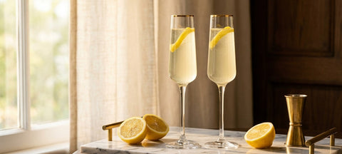 Two French 75 cocktails with lemon twists in gold-rimmed champagne flutes on a marble tray beside cut lemons
