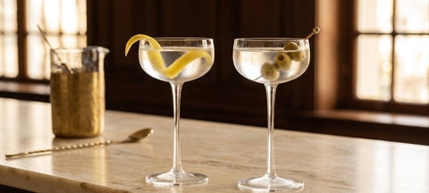 Two classic dry martinis with a lemon twist and olives in Nick and Nora coupe glasses on a marble bar in warm window light