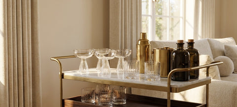 A brass and marble bar cart in a living room stocked with crystal coupes, ribbed lowball glasses, a shaker and an ice bucket