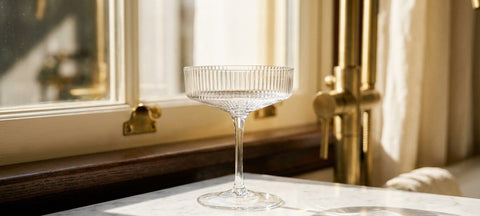 A single ribbed crystal coupe glass catching bright window light on a marble surface