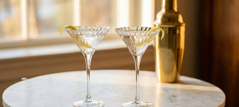 Two seashell-ribbed V-shaped martini glasses with dry martinis and a lemon twist on a marble table beside a brass shaker