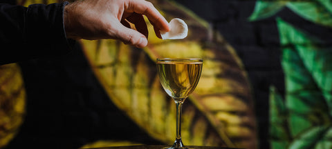 A hand dropping a garnish into a single Nick and Nora glass on a dark marble bar