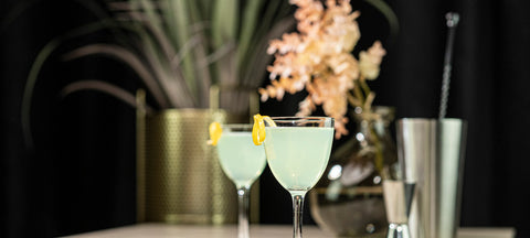 Two Nick and Nora glasses with a pale green cocktail in front of a cocktail shaker and bar spoon
