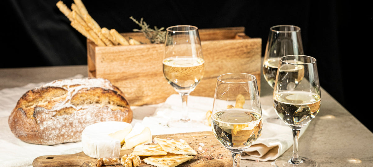 Stemmed tasting glasses of white wine with a loaf of bread, cheese and grissini on a linen table